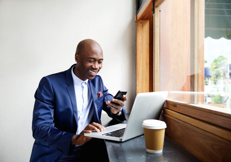51498038 - portrait of happy african businessman using phone while working on laptop in a restaurant